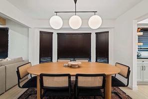 Un comedor con una mesa redonda de madera, cuatro sillas, una lámpara central y persianas oscuras en las ventanas.