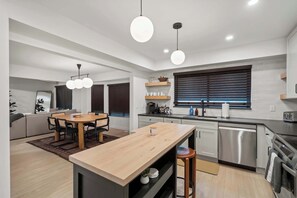 Eine moderne Küche mit einer zentralen Kochinsel, Pendelleuchten und einem Essbereich mit Sofa und Tisch.