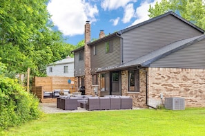 Una casa de dos pisos con fachada de ladrillo y techo gris, un patio con mobiliario exterior y una chimenea.