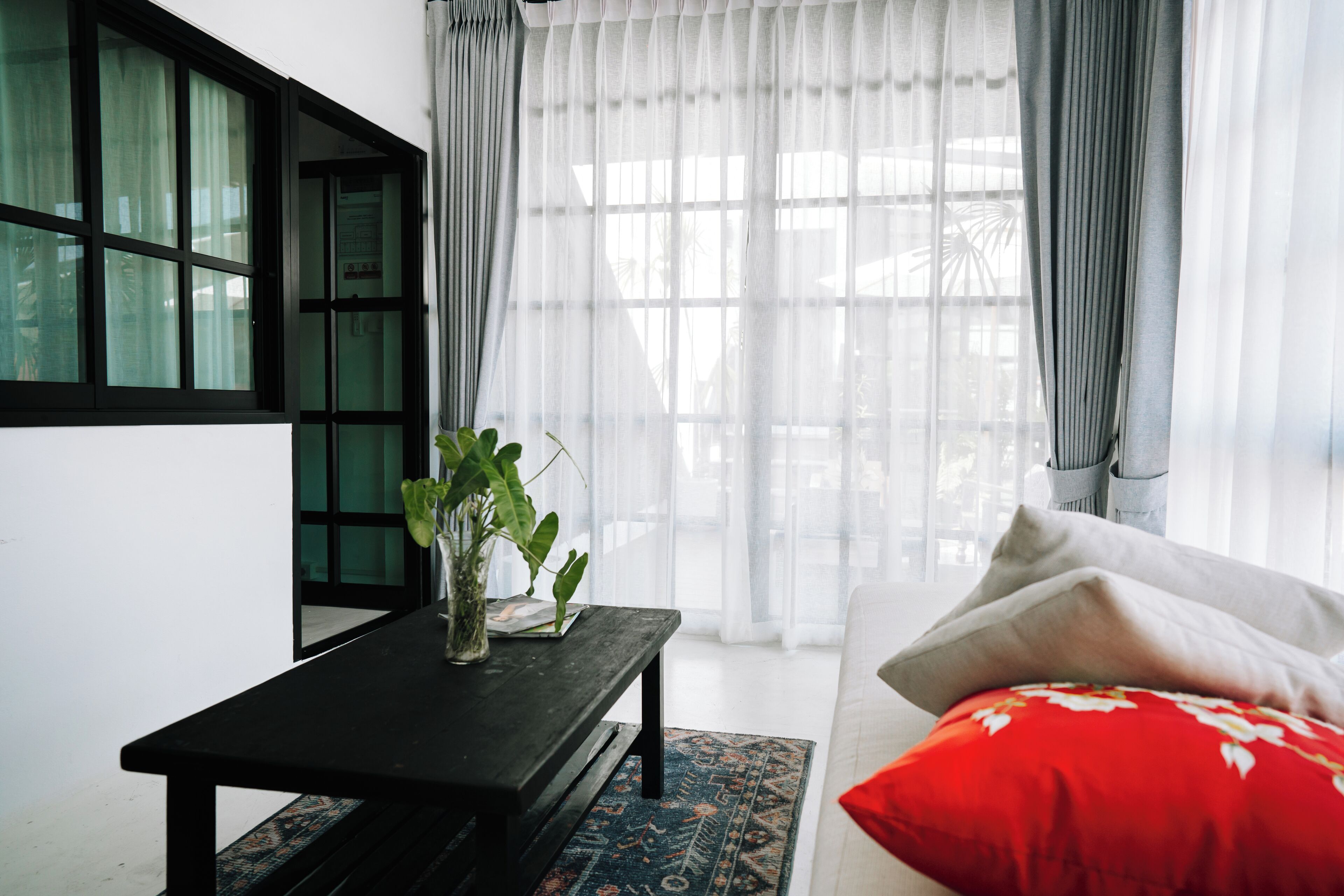 Un salon moderne avec une table basse noire, un canapé blanc agrémenté d’un coussin rouge et une grande fenêtre ornée de rideaux légers.