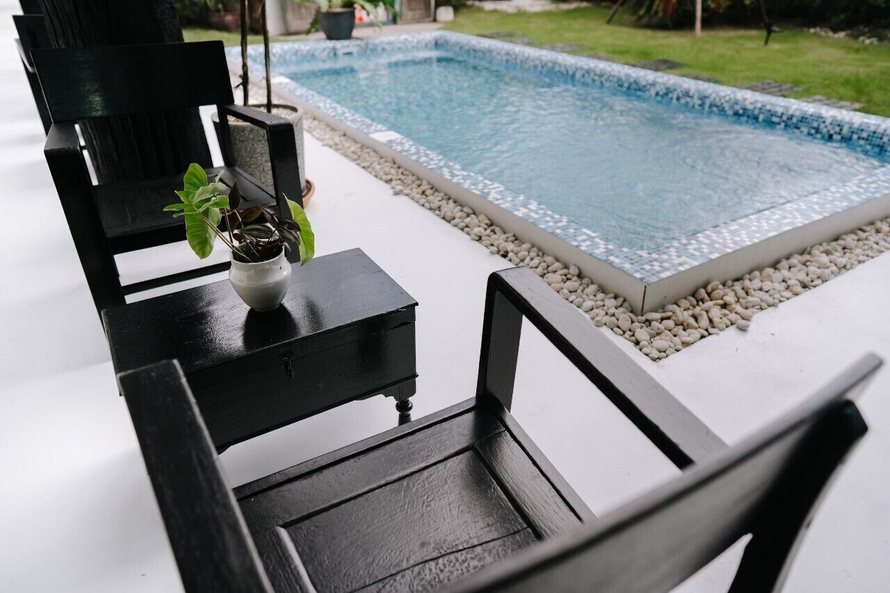 A poolside area with a black chair, a small table with a potted plant, and a tiled pool surrounded by pebbles.