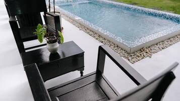 A poolside area with a black chair, a small table with a potted plant, and a tiled pool surrounded by pebbles.