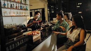 Zona de bar con estantes llenos de botellas y vasos, un mostrador con comida, y un grupo de personas sentadas.