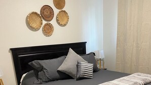 Una cama bien hecha con un cabecero oscuro, ropa de cama gris y una manta de rayas grises y blancas. Hay dos mesitas de noche con lámparas y una cesta fijada a la pared.