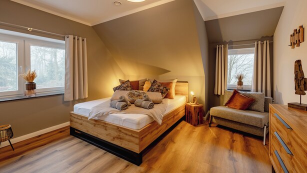 A bedroom with a wooden bed, a sofa, a nightstand, and a window with curtains. - Hünzinger Dorfschmiede - Holiday apartment Specht (Walsrode)