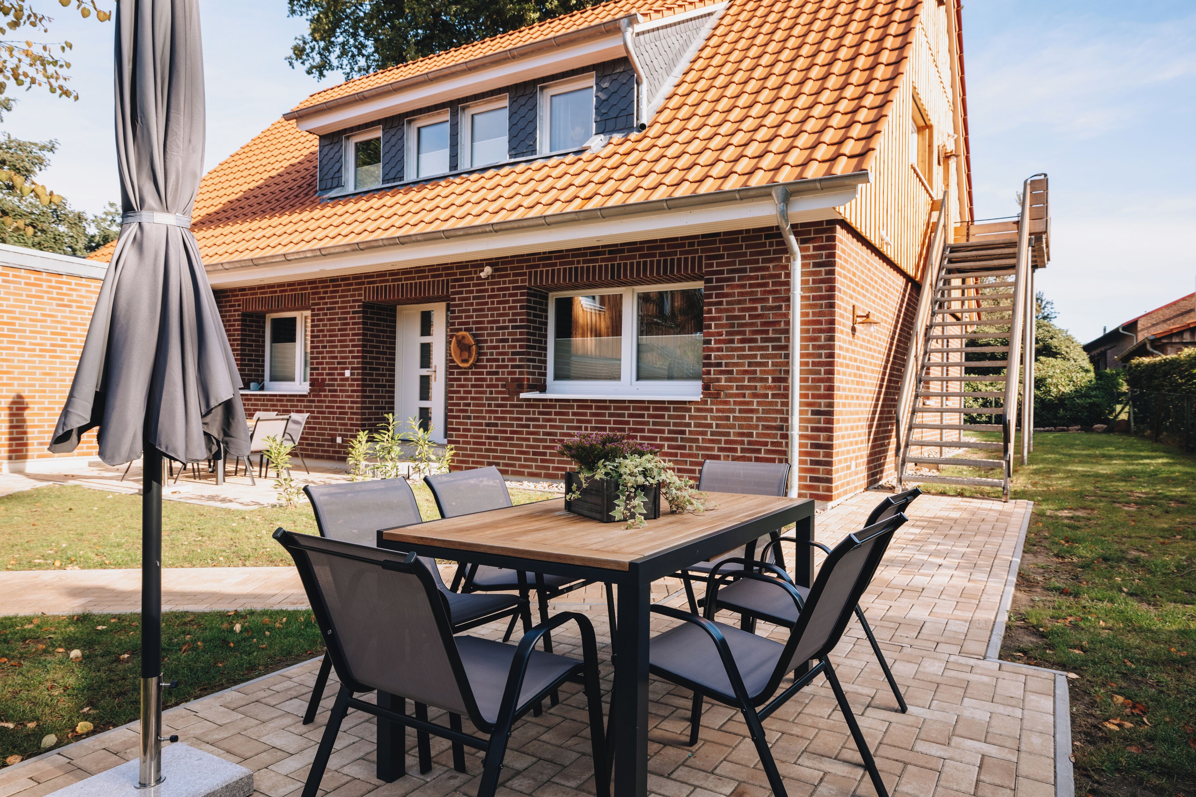 Eine Terrasse mit Holztisch und -stühlen, einem grauen Sonnenschirm und einem Haus mit Ziegeldach.