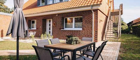 Eine Terrasse mit Holztisch und -stühlen, einem grauen Sonnenschirm und einem Haus mit Ziegeldach.