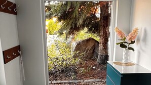 Une pièce donnant sur un jardin, avec un plancher en bois, un tapis vert, une armoire bleue et un vase blanc rempli de fleurs.