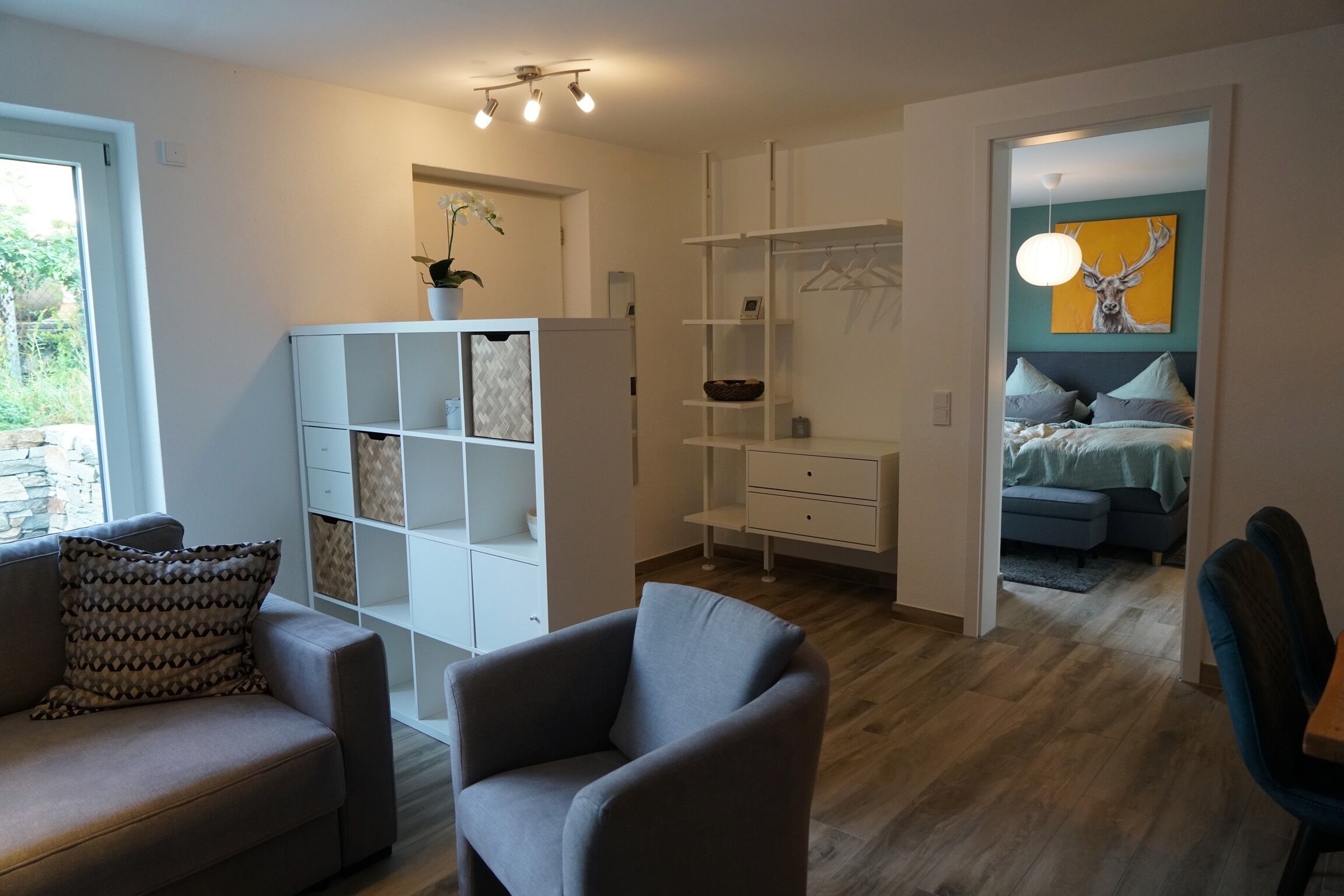 Een moderne woonkamer met een bank, fauteuil en een ingebouwde boekenkast. Op de achtergrond staat een bed met een blauwe hoofdbord, en aan de muur hangt een schilderij van een hert.