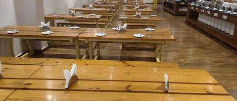 A cafeteria with wooden tables and chairs, each with a white plate and a silver spoon.