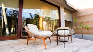 Un espacio exterior moderno con sillas de mimbre, una mesa, amplios ventanales de vidrio y piso de madera.