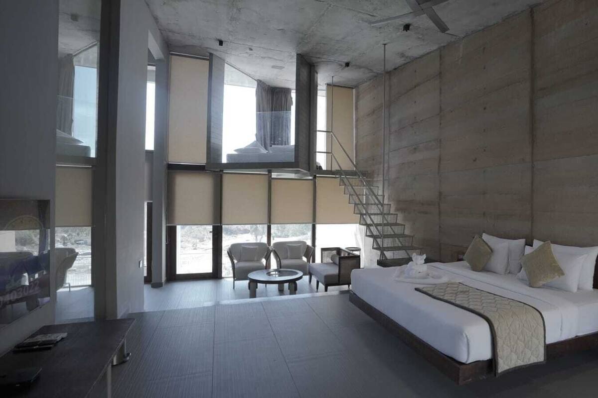 Una habitación de hotel moderna con una cama grande, un área de descanso con dos sillones, una mesita y una escalera que lleva a un nivel superior.