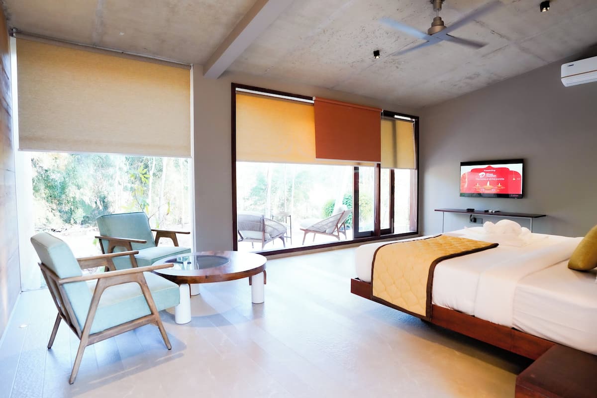 Una habitación de hotel moderna con cama, televisor, zona de estar con sillas y un ventanal que ofrece vistas al exterior.
