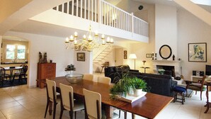 Un amplio salón con una mesa de comedor, sillas, una araña, una escalera y una chimenea.
