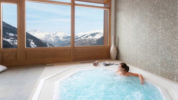 A spa room with a hot tub, wooden windows, and a view of snow-capped mountains.