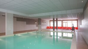 A swimming pool with a tiled floor and walls, a ceiling with recessed lighting, and a row of red chairs along the wall.