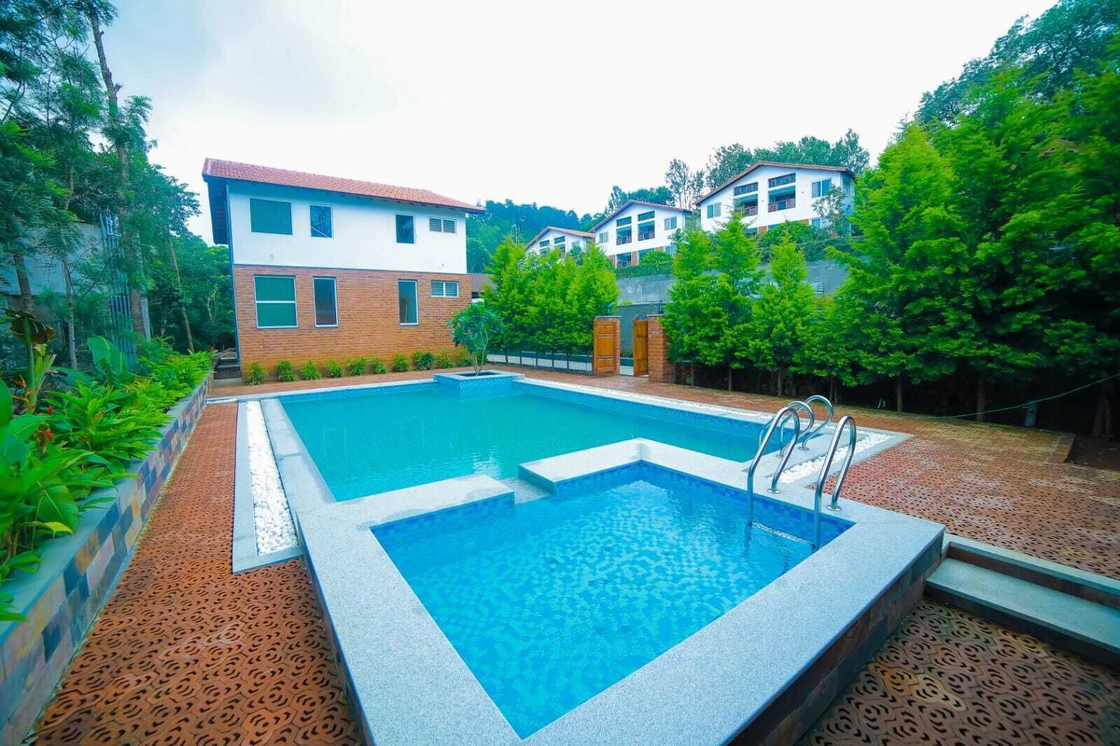 A rectangular swimming pool with a tiled deck, surrounded by lush greenery and a brick building.