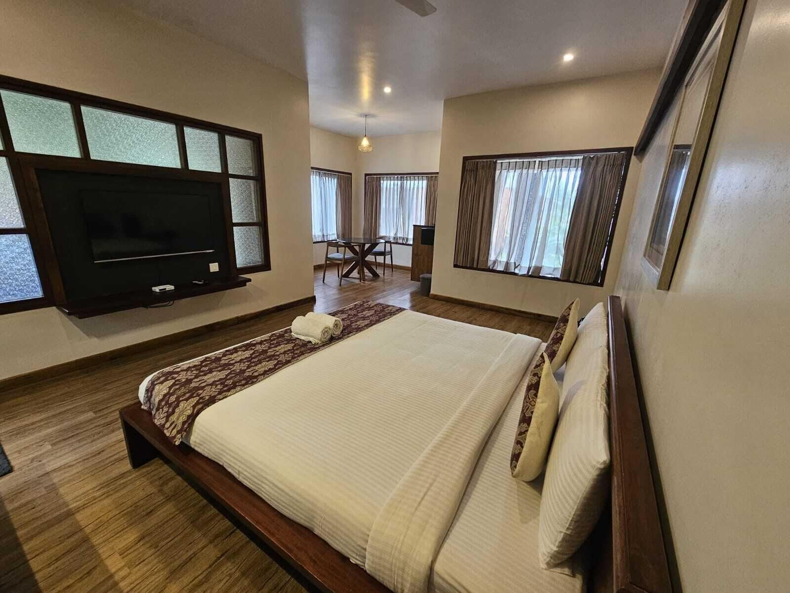 A hotel room with a large bed, a flat-screen TV mounted on the wall, a dining area with a table and chairs, and large windows with curtains.