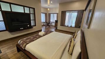 A hotel room with a large bed, a flat-screen TV mounted on the wall, a dining area with a table and chairs, and large windows with curtains.