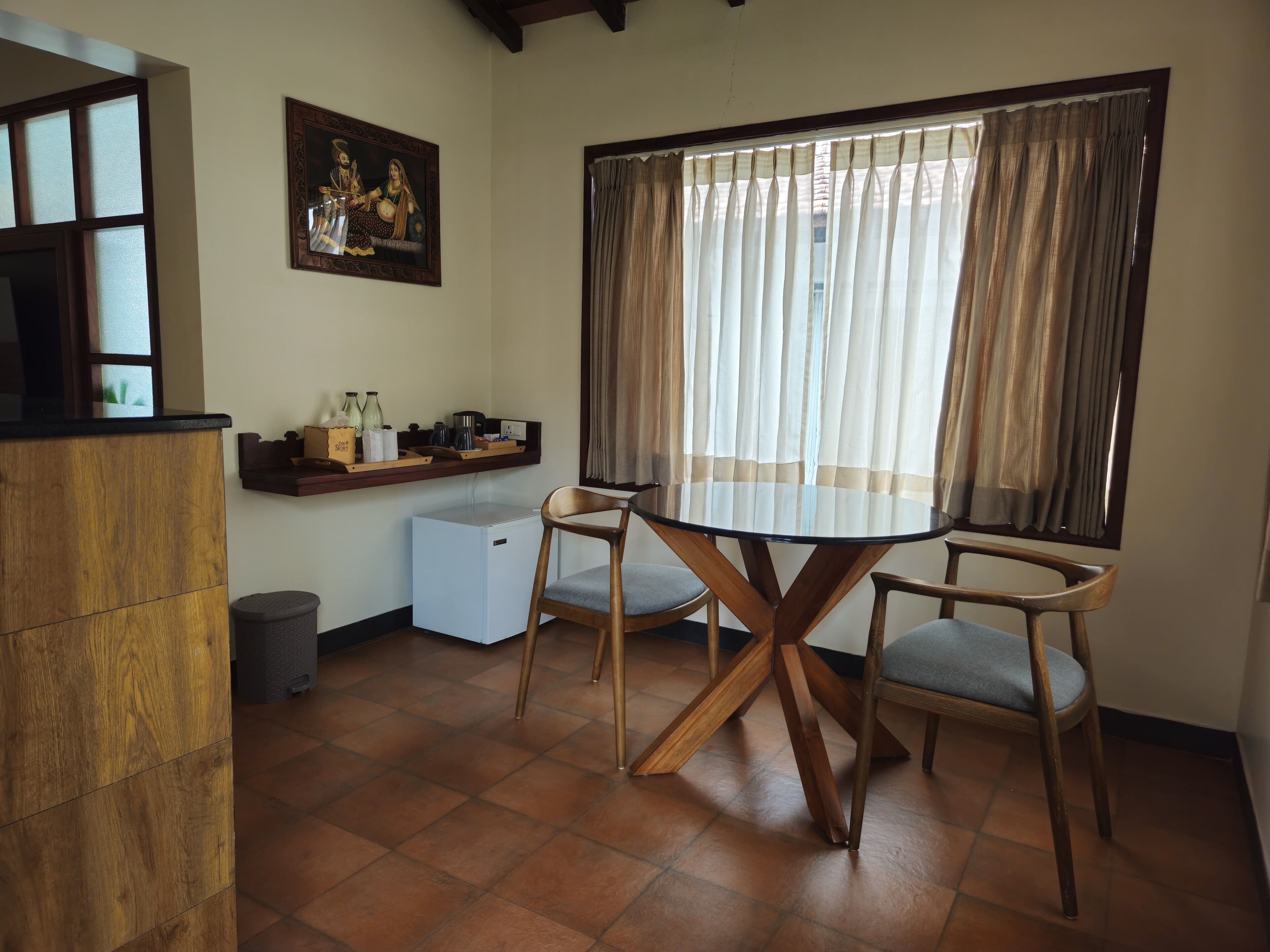 Un coin repas comprenant une table ronde et deux chaises, un petit réfrigérateur, une étagère avec des ustensiles de cuisine et une fenêtre avec des rideaux.
