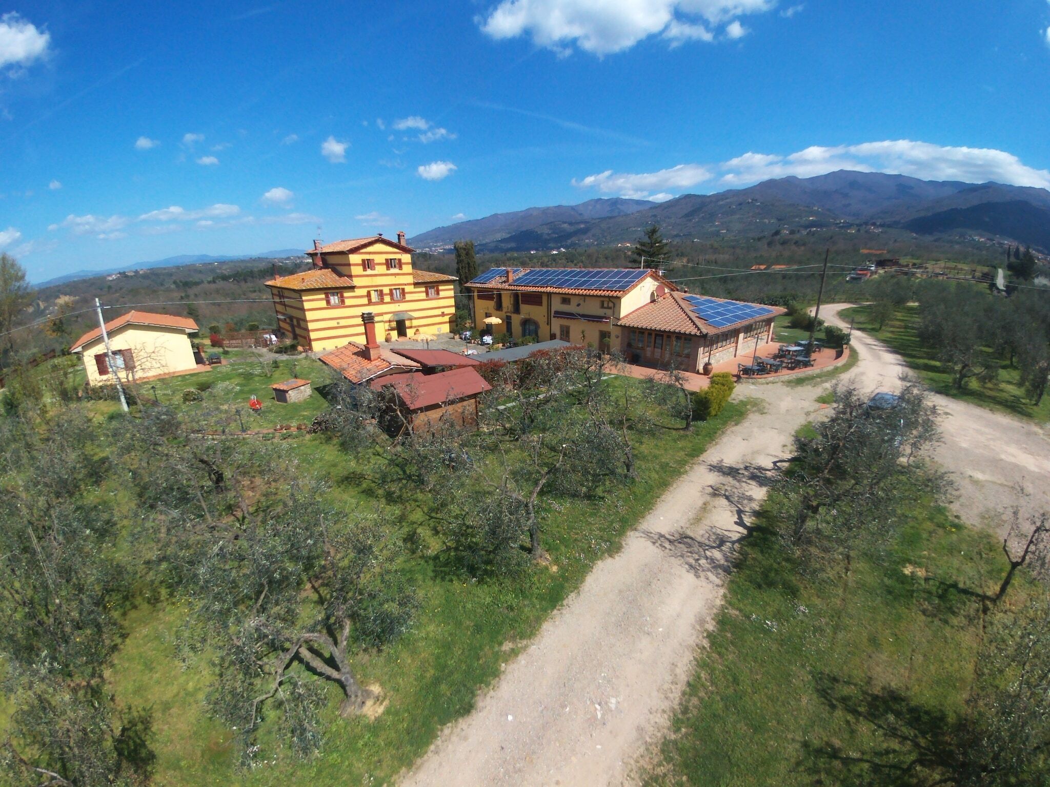 A large yellow building with multiple rooms and a solar panel roof, surrounded by green fields and mountains.
