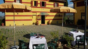 Eine Außenpatio mit Tischen und Stühlen, ein gelbes Gebäude mit roten Streifen und Blick auf die Landschaft.