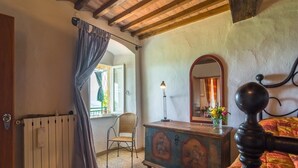 A bedroom with a wooden ceiling, a radiator, a chair, a dresser with religious paintings, a mirror, and a bed with a headboard.