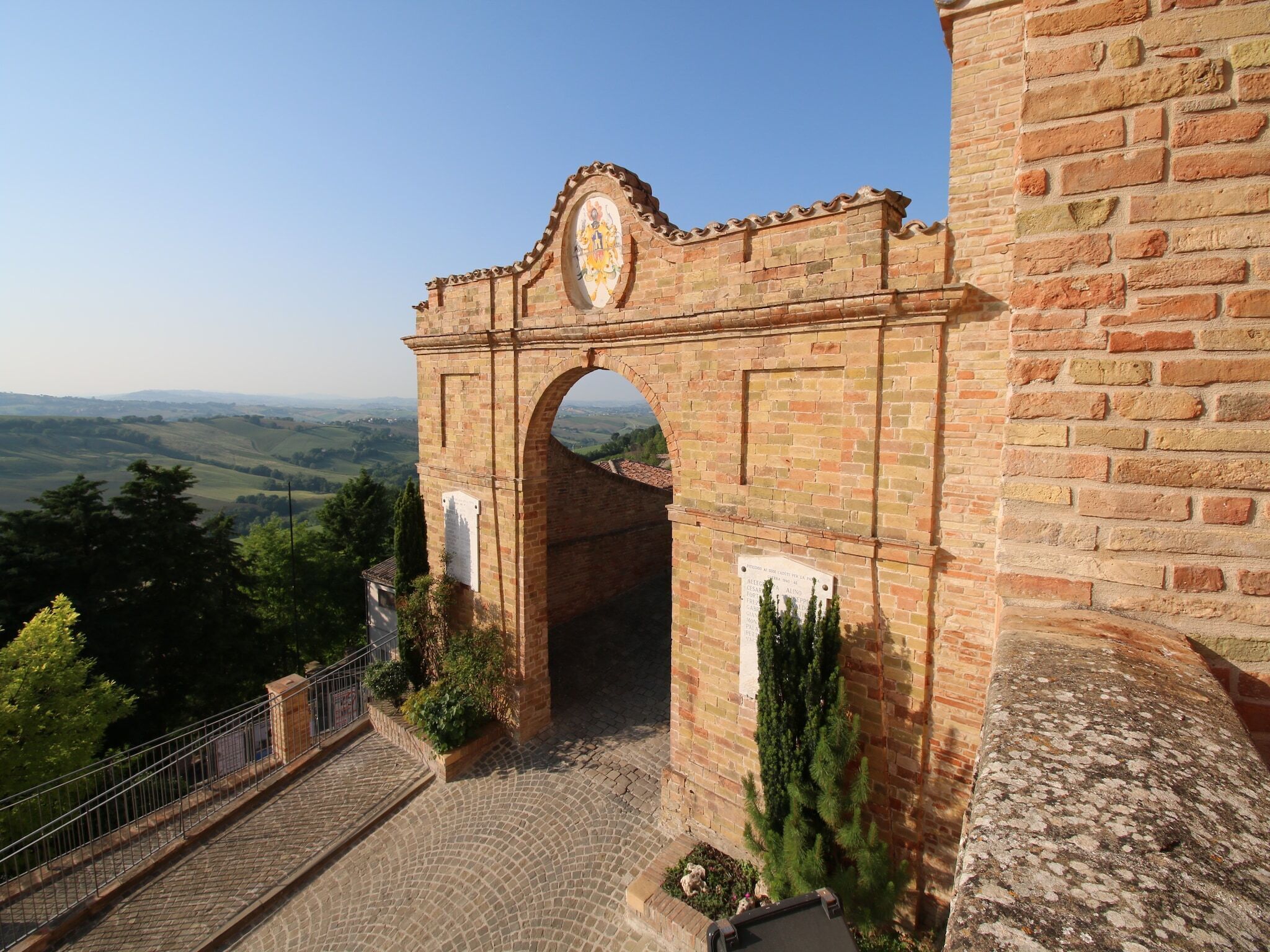 Un arco de piedra con una placa, que conduce a un área pavimentada con barandilla metálica y vistas al campo.