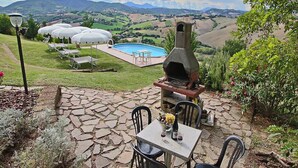 An outdoor patio with stone flooring, a table with chairs, a barbecue grill, and a swimming pool in the background. - Apartment in Piticchio with Pool and BBQ (Arcevia)