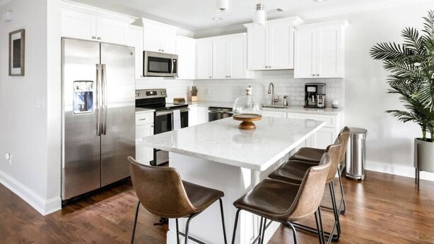 A modern kitchen with white cabinetry, stainless steel appliances, a marble island, and a potted plant. - Garden Home at Liberty Estate (Savannah)