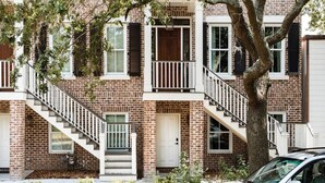 A two-story brick building with a white front door, a small front porch, and a staircase leading to the second floor. - Garden Home at Liberty Estate (Savannah)