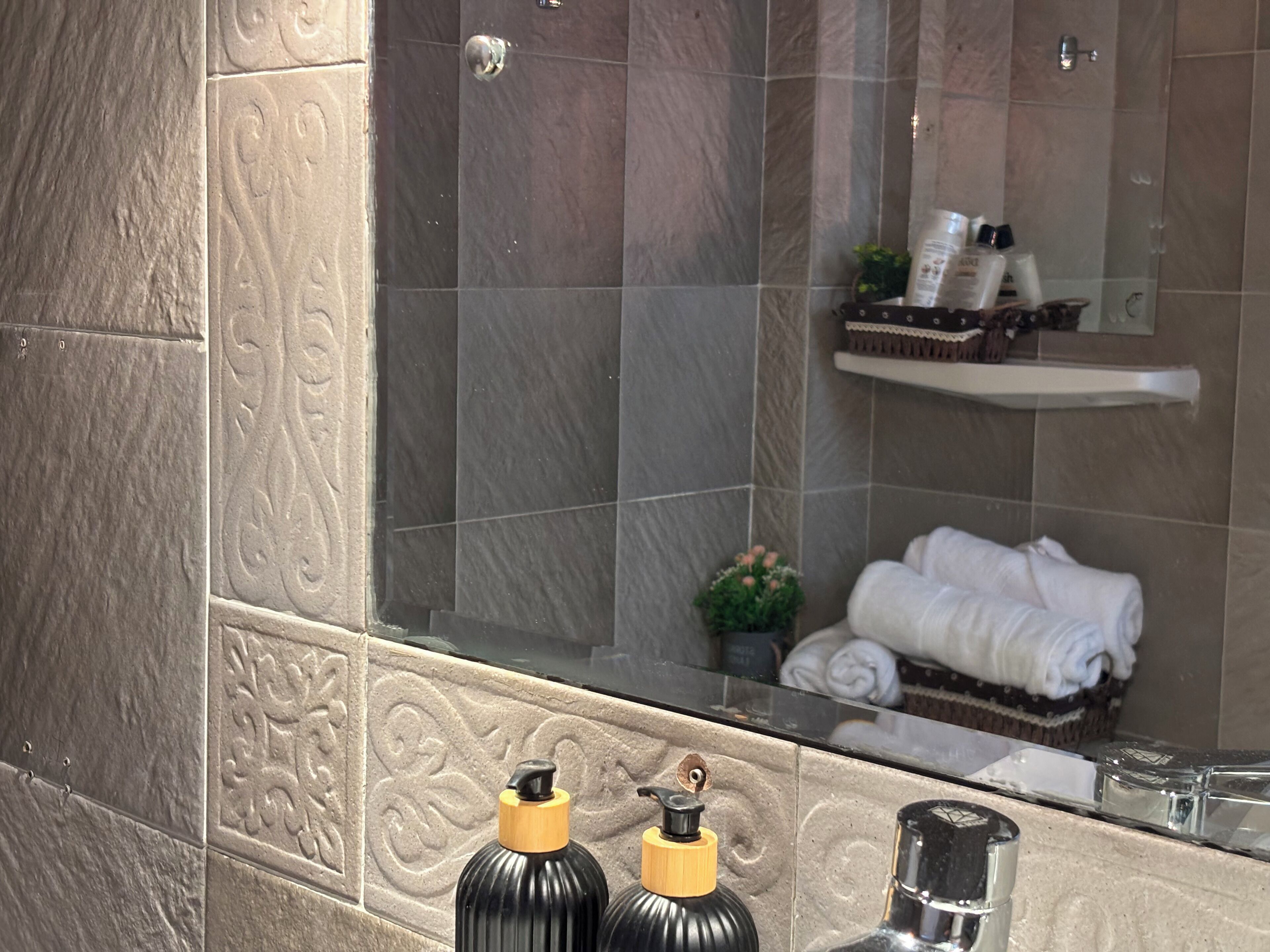 A bathroom with a mirror reflecting a towel rack and toiletries. The walls are tiled with a stone-like pattern.