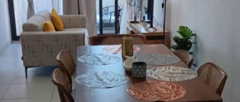 A dining area with a table set for four, surrounded by wicker chairs, with a sofa and curtains in the background.