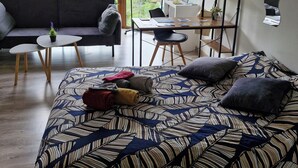A room with a large bed, a sofa, a small table, and a desk. There are large windows with a view of houses and trees. - Lovely studio 15min walk from Epinal town center (Chantraine)