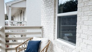 Un balcon avec des meubles en osier et des coussins blancs, un mur en briques blanches et une fenêtre.
