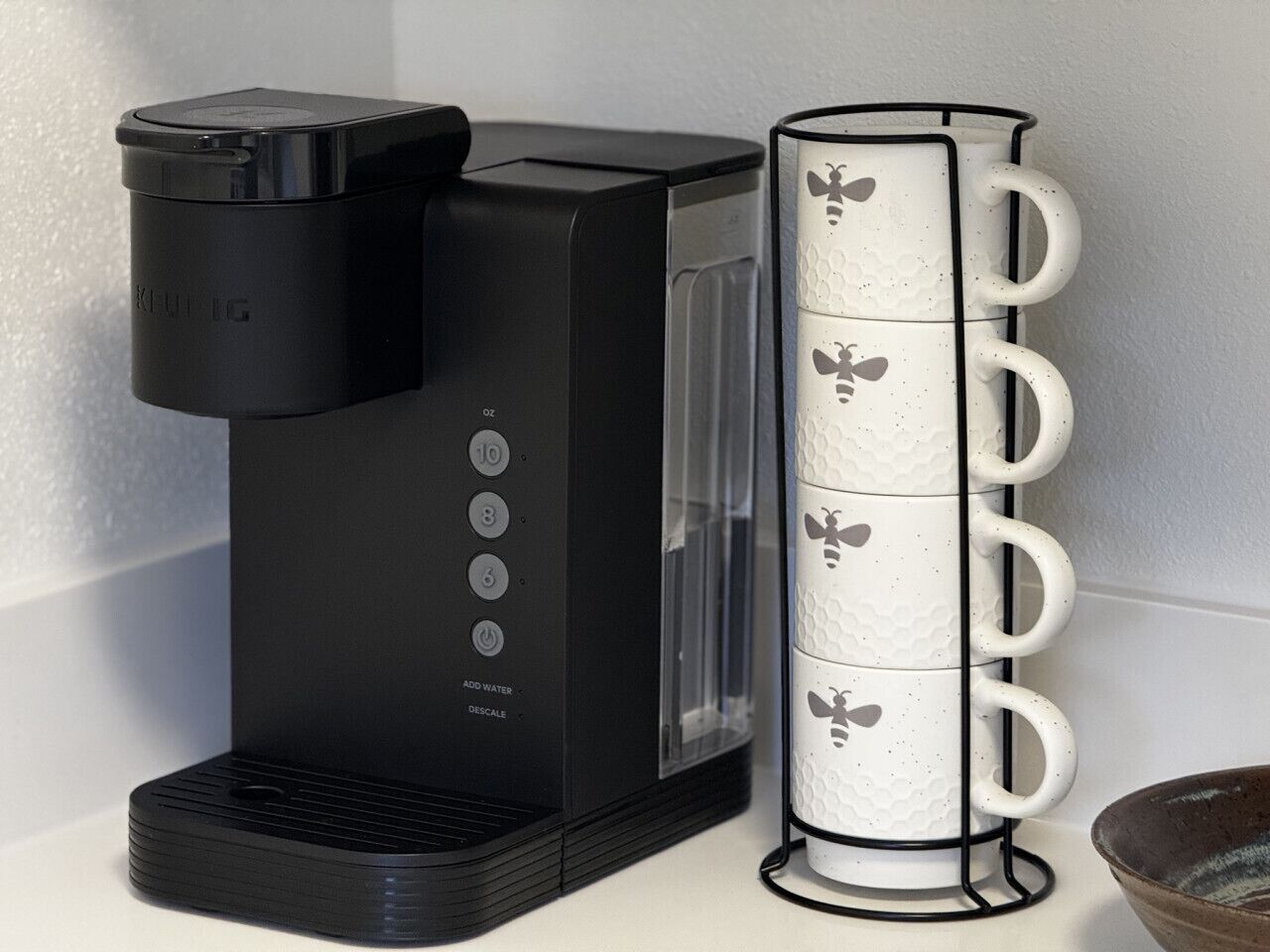 Una cafetera con depósito de agua y una pila de tazas blancas con diseños de abejas.