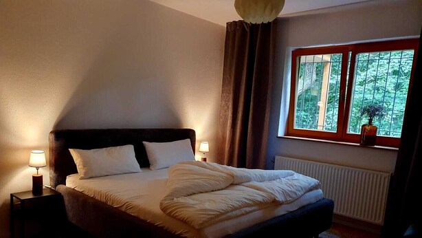 Une chambre à coucher avec un lit, des tables de chevet, une fenêtre donnant sur des arbres et un radiateur.