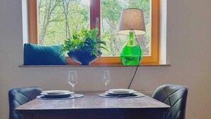 Un coin repas avec une table dressée pour deux personnes, une fenêtre donnant sur des arbres, un coussin bleu et une lampe verte.