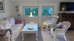 Uma sala de estar acolhedora com um sofá floral, uma mesa branca com velas, uma poltrona de vime e uma TV na parede.