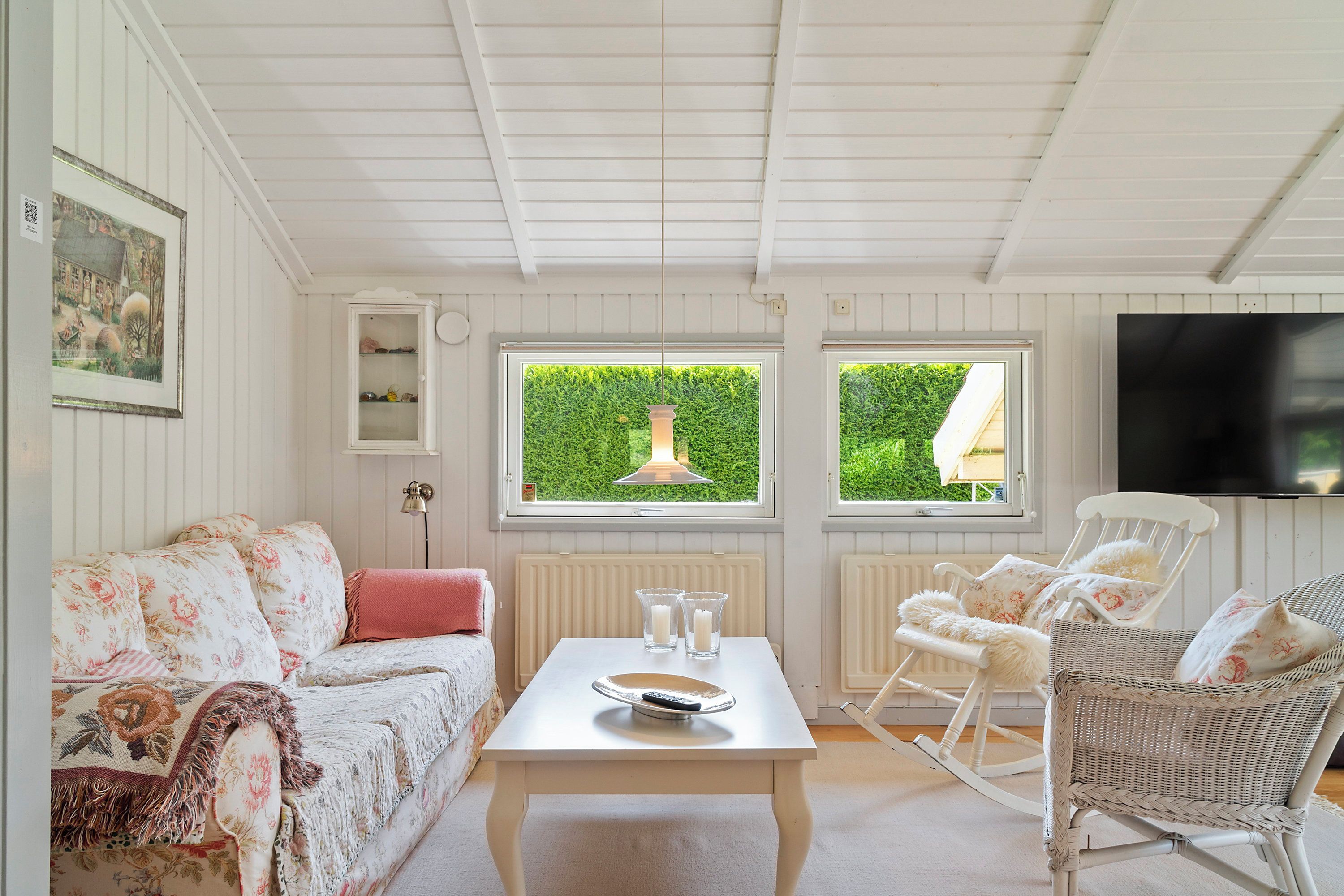 A cozy living room with a white sofa, a coffee table, and wicker chairs. There are windows with a view of greenery, a wall-mounted TV, and a framed picture on the wall.