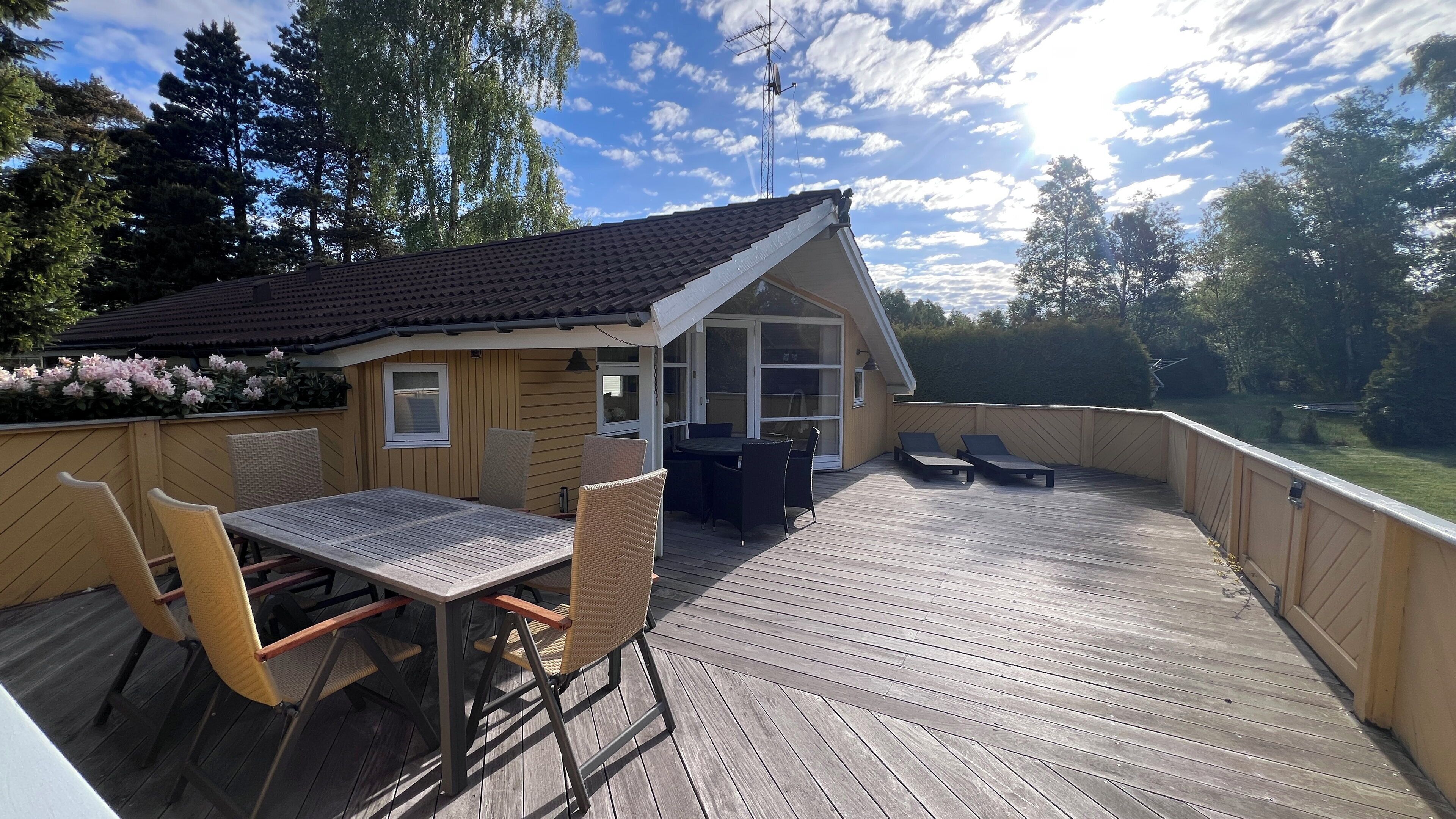 A wooden deck with a table and chairs, a small building with a sloped roof, and a view of trees and a body of water.