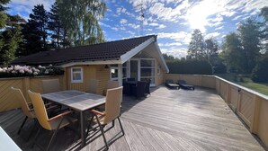 A wooden deck with a table and chairs, a small building with a sloped roof, and a view of trees and a body of water. - Super cozy holiday home in Marielyst (Væggerløse)