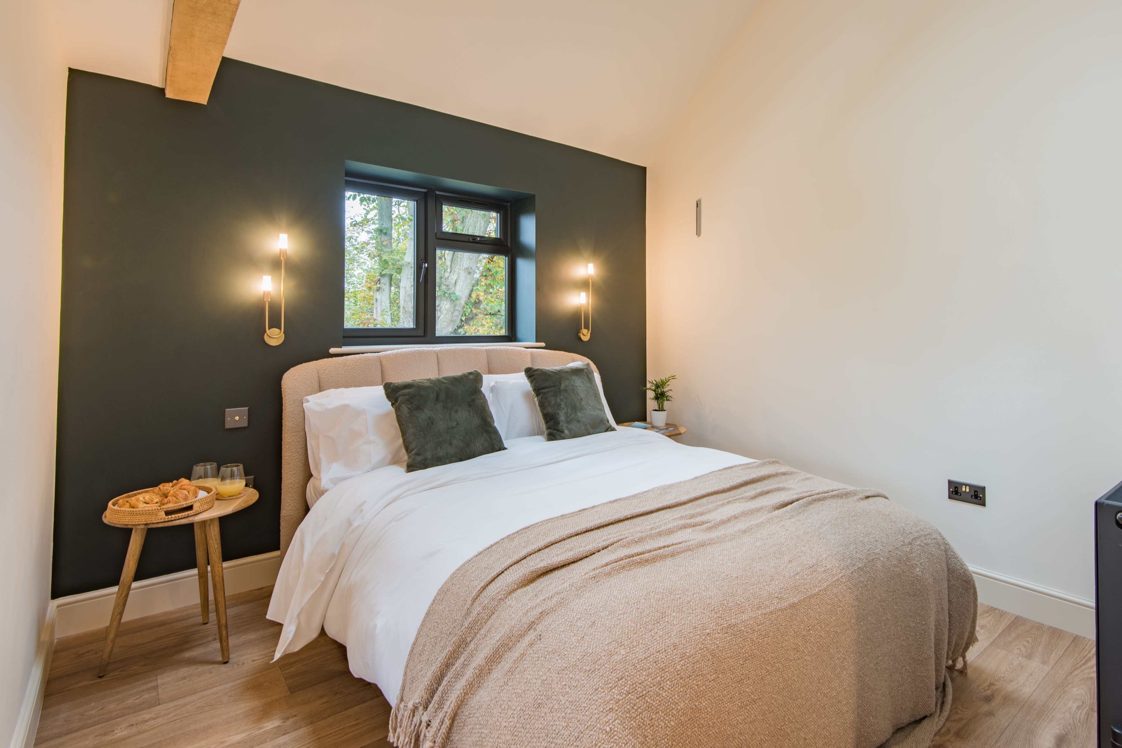 A bedroom with a large bed, two wall-mounted lights, a small table with a basket, and a window with a view of trees.