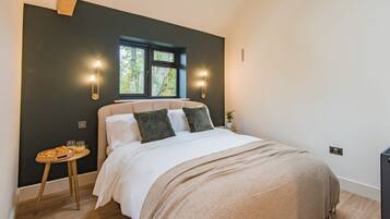 A bedroom with a large bed, two wall-mounted lights, a small table with a basket, and a window with a view of trees.