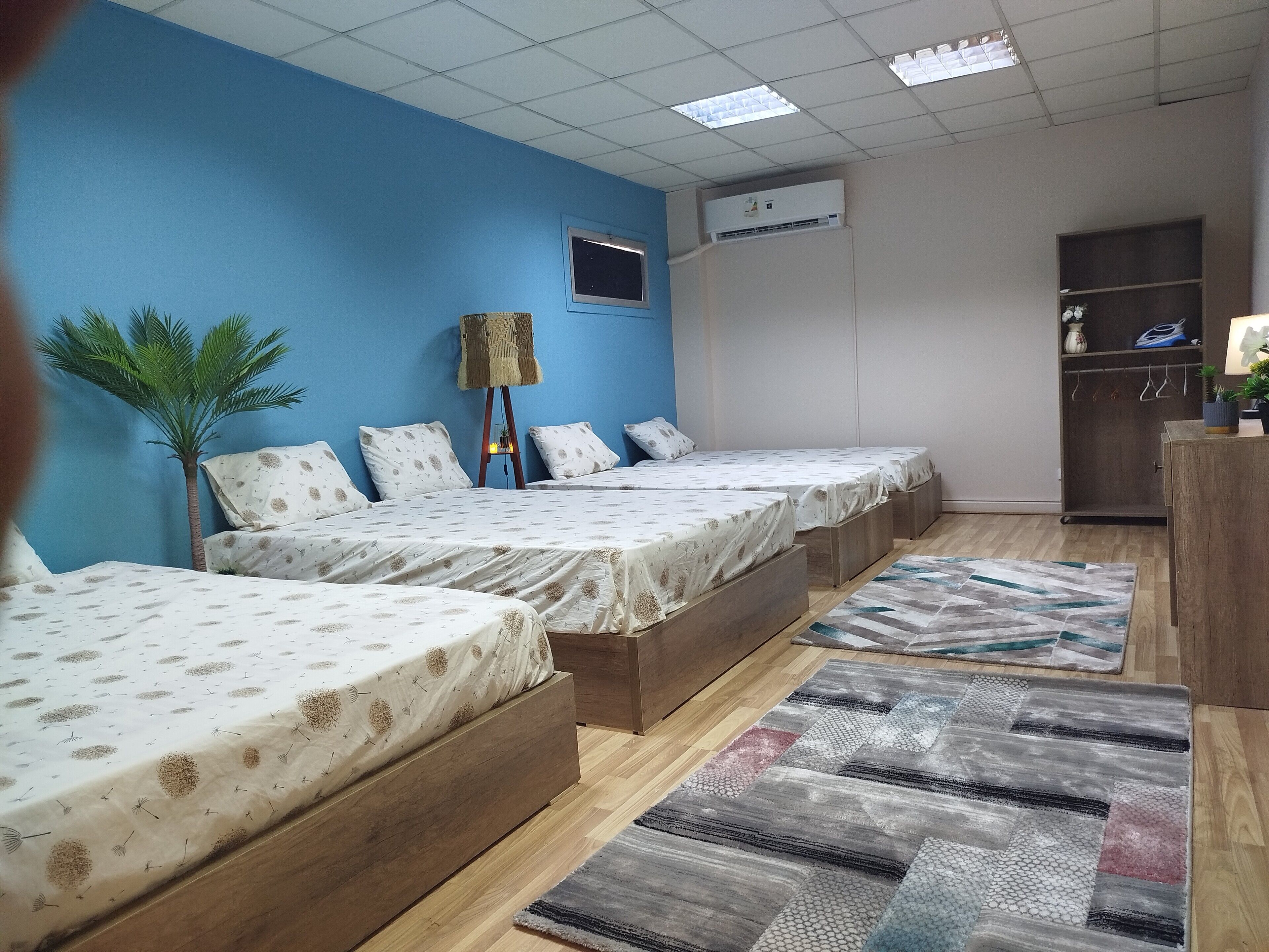 Habitación de hotel con tres camas individuales, piso de madera, pared de acento azul, unidad de aire acondicionado, un escritorio pequeño con lámpara y un armario.