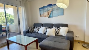 A modern living room with a grey sofa, a glass coffee table, and a view of a balcony with outdoor furniture.