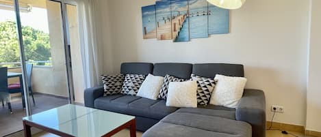 Una sala de estar moderna con un sofá gris, una mesa de centro de vidrio y vistas a un balcón con mobiliario exterior.
