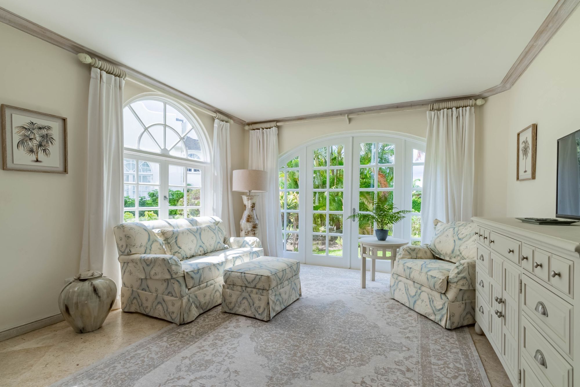 A spacious living room with a large arched window, a sofa set, a side table with a lamp, and a cabinet.