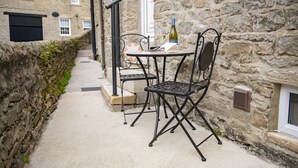 A stone building with a narrow alleyway, a metal table with chairs, and a bottle of wine.