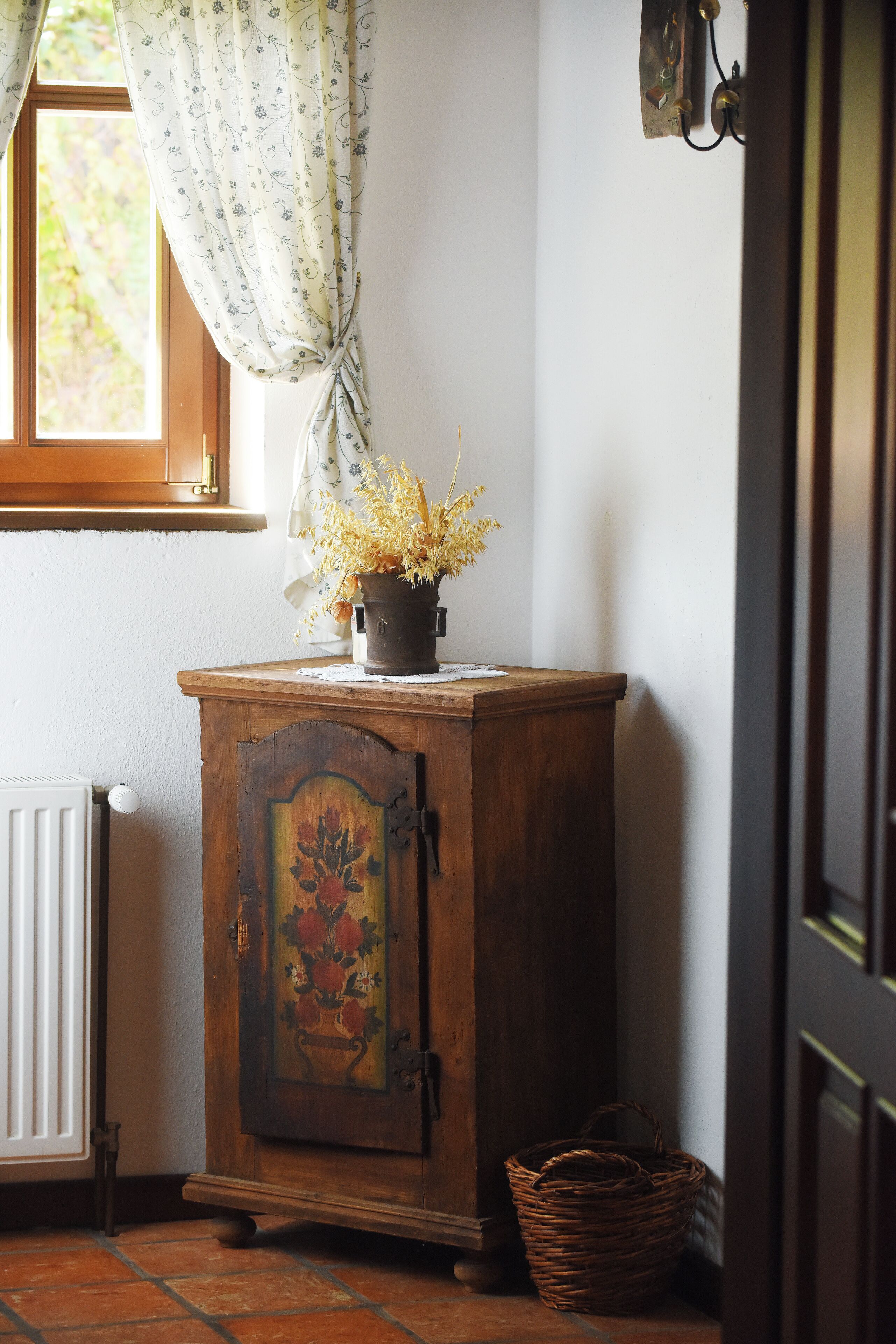 A wooden cabinet with a floral design, a potted plant, a wicker basket, and a radiator.